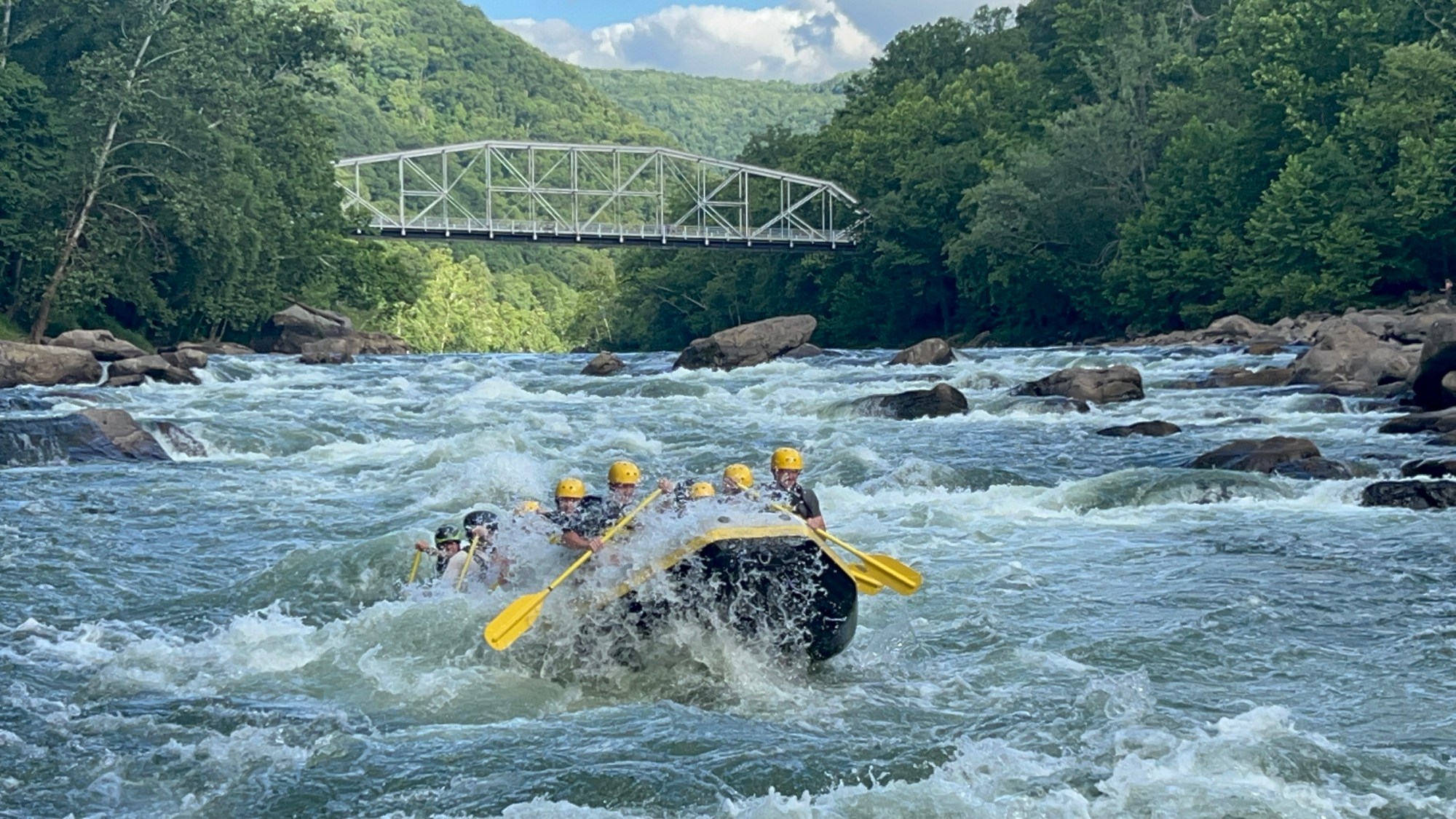 Group whitewater rafting under a bridge surrounded by forest.