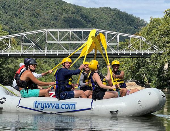 Group in helmets and life vests rafting under a bridge on a river.