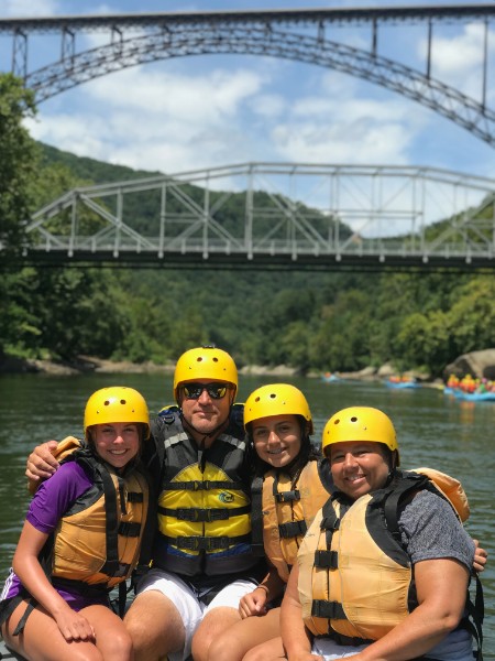 Four people in helmets and life vests on a river, with bridges and trees in the background.