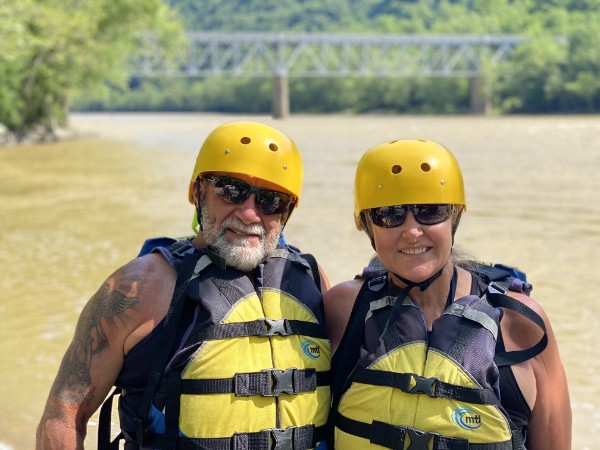 Two people in helmets and life jackets stand by a river with a bridge in the background.