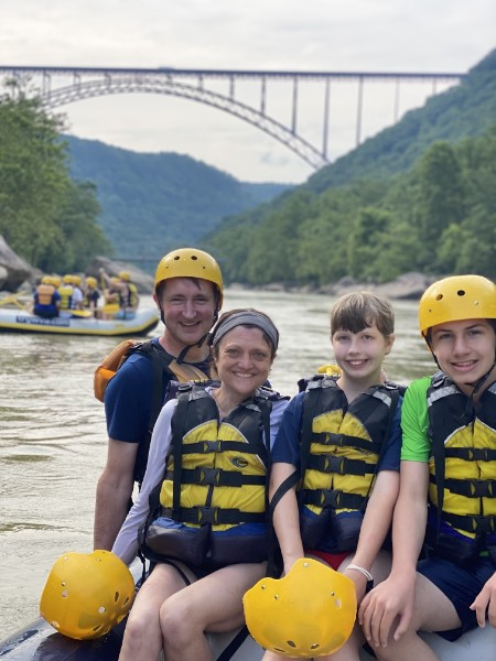 Family in helmets and life jackets rafting on a river with a bridge in the background.