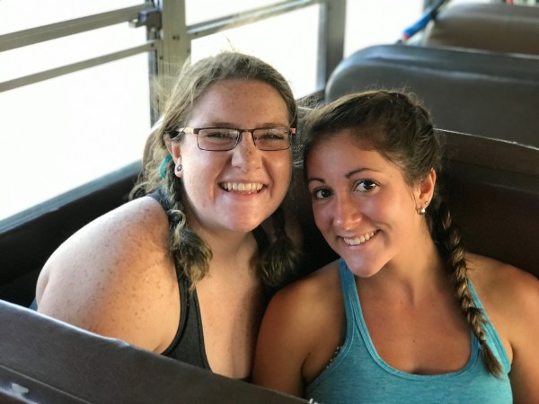 Two women smiling and sitting together on a bus with sunlight streaming in.