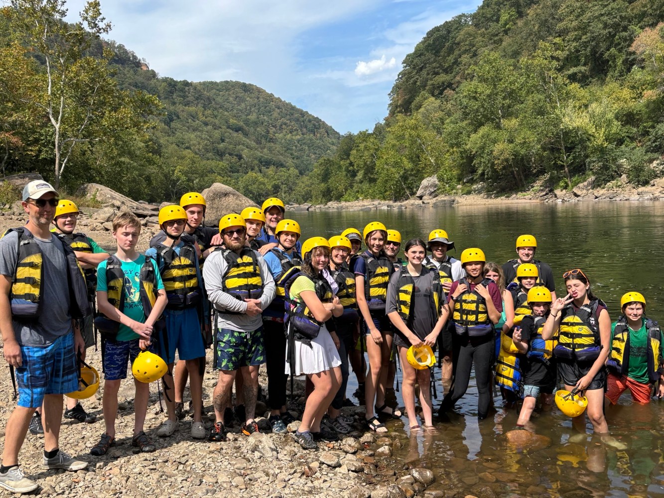 Group in helmets and life vests by a river, set against a forested hill background.