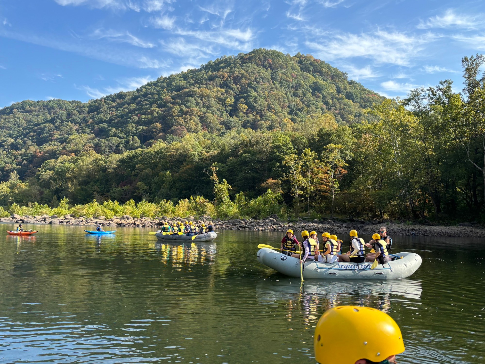 Rafting group in helmets on a river with a forested hill in the background.
