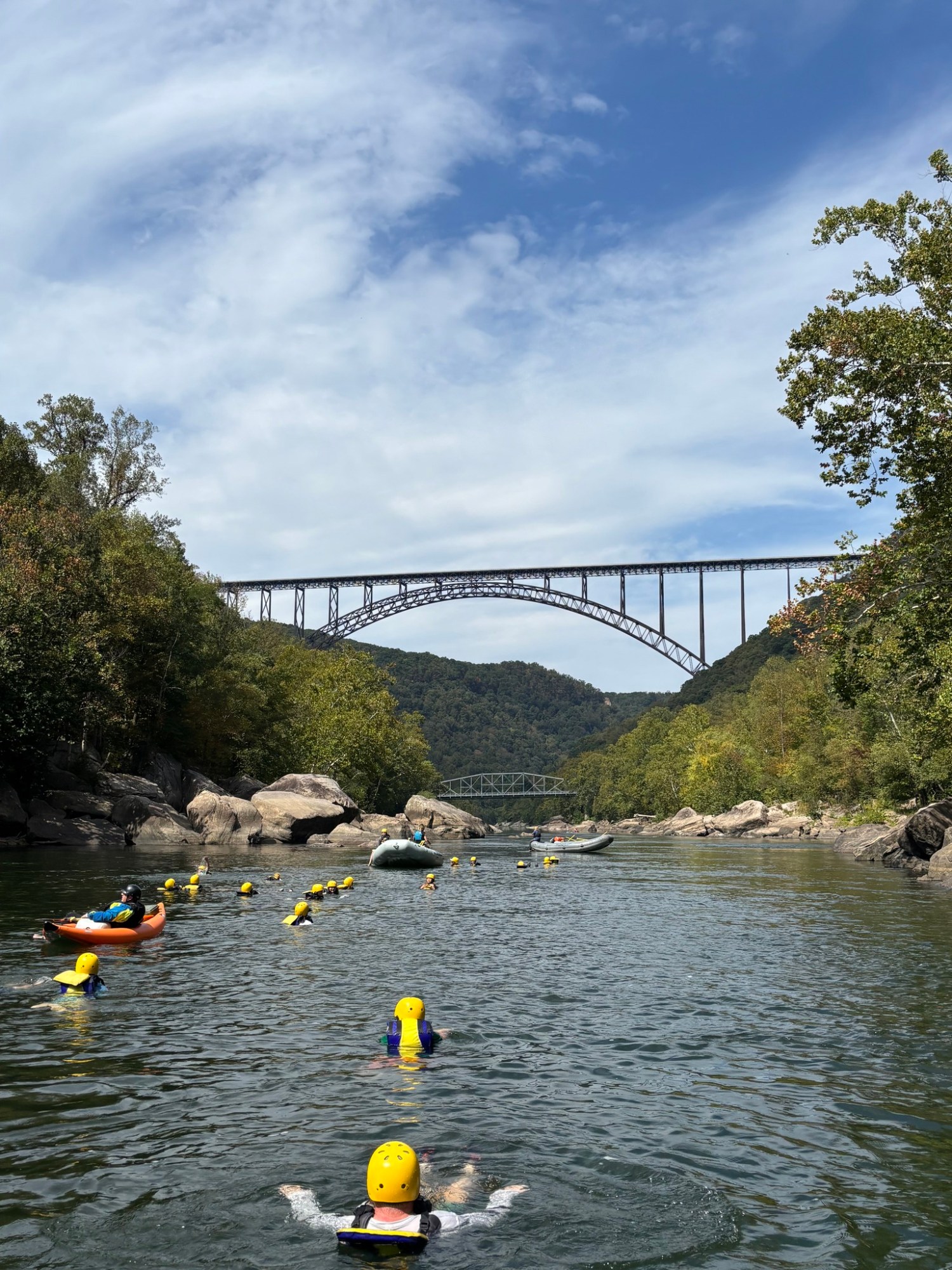 People in helmets floating in a river with a bridge overhead and trees surrounding.