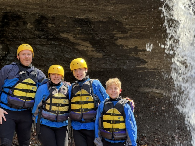 Four people wearing helmets and life jackets stand near a waterfall.