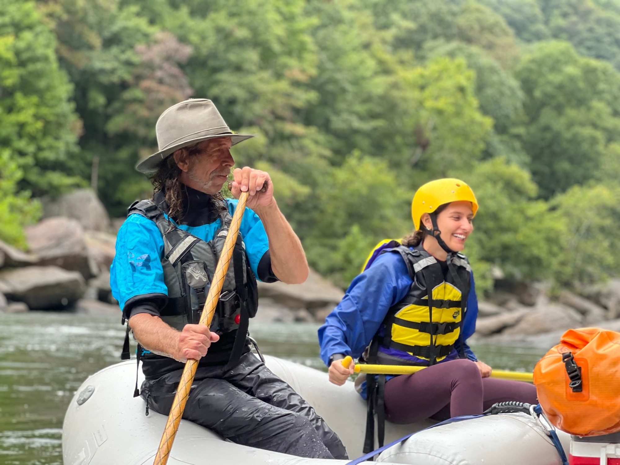 Two people in helmets and life vests rafting on a river surrounded by trees.