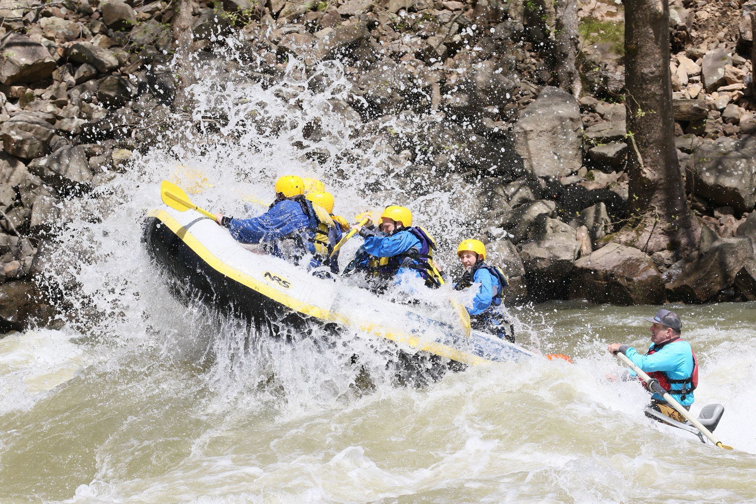 Group of people white-water rafting in a river, wearing helmets and life jackets, surrounded by splashing water.