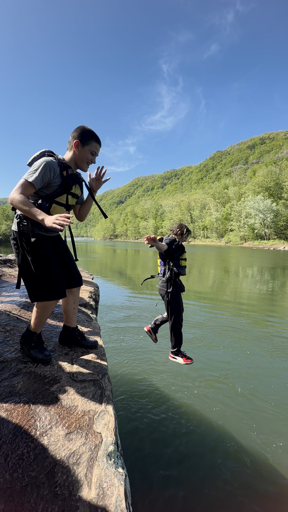 Two people in life vests jump into a lake from a rocky ledge.