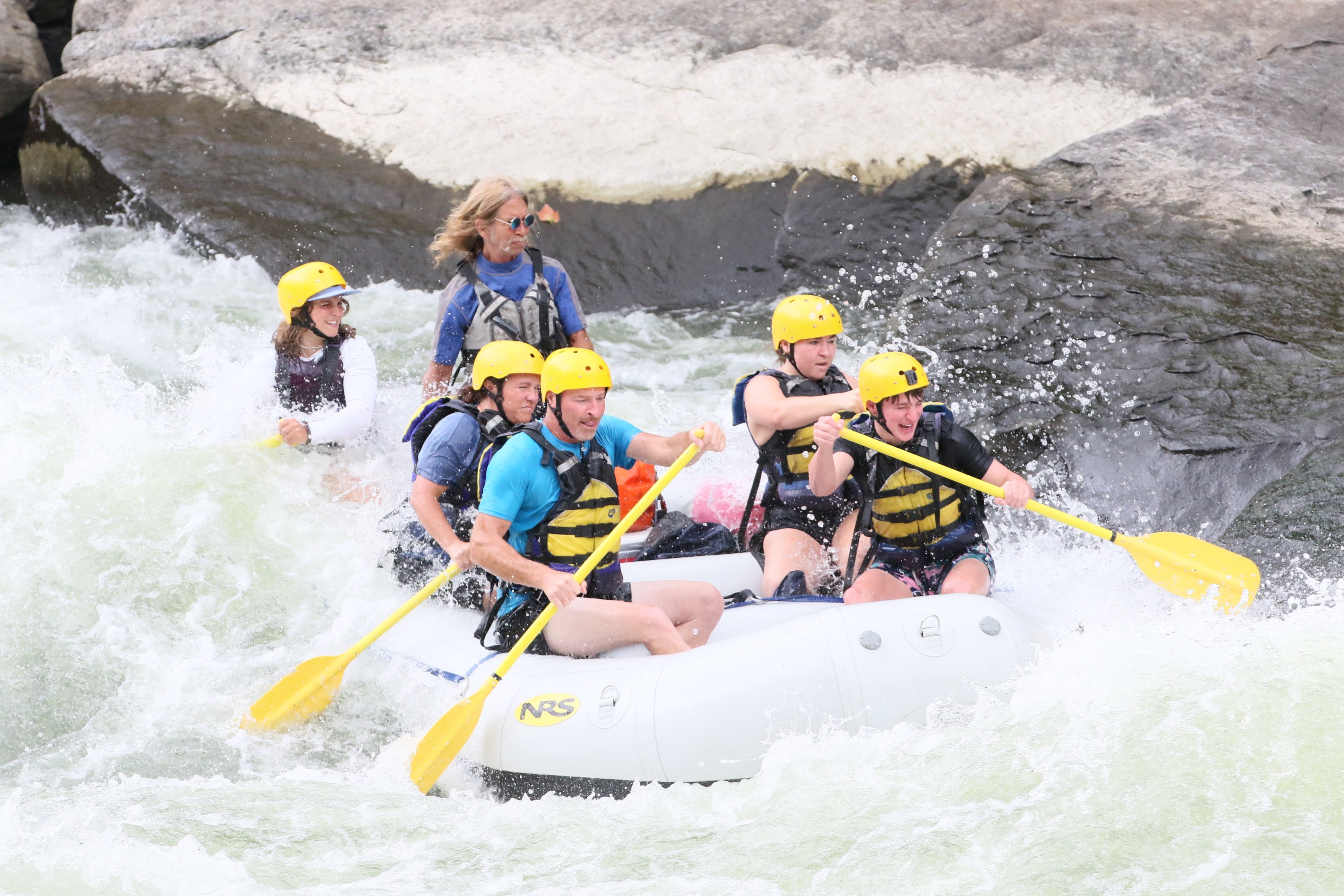 Group rafting in whitewater, wearing helmets and life jackets, paddling vigorously.