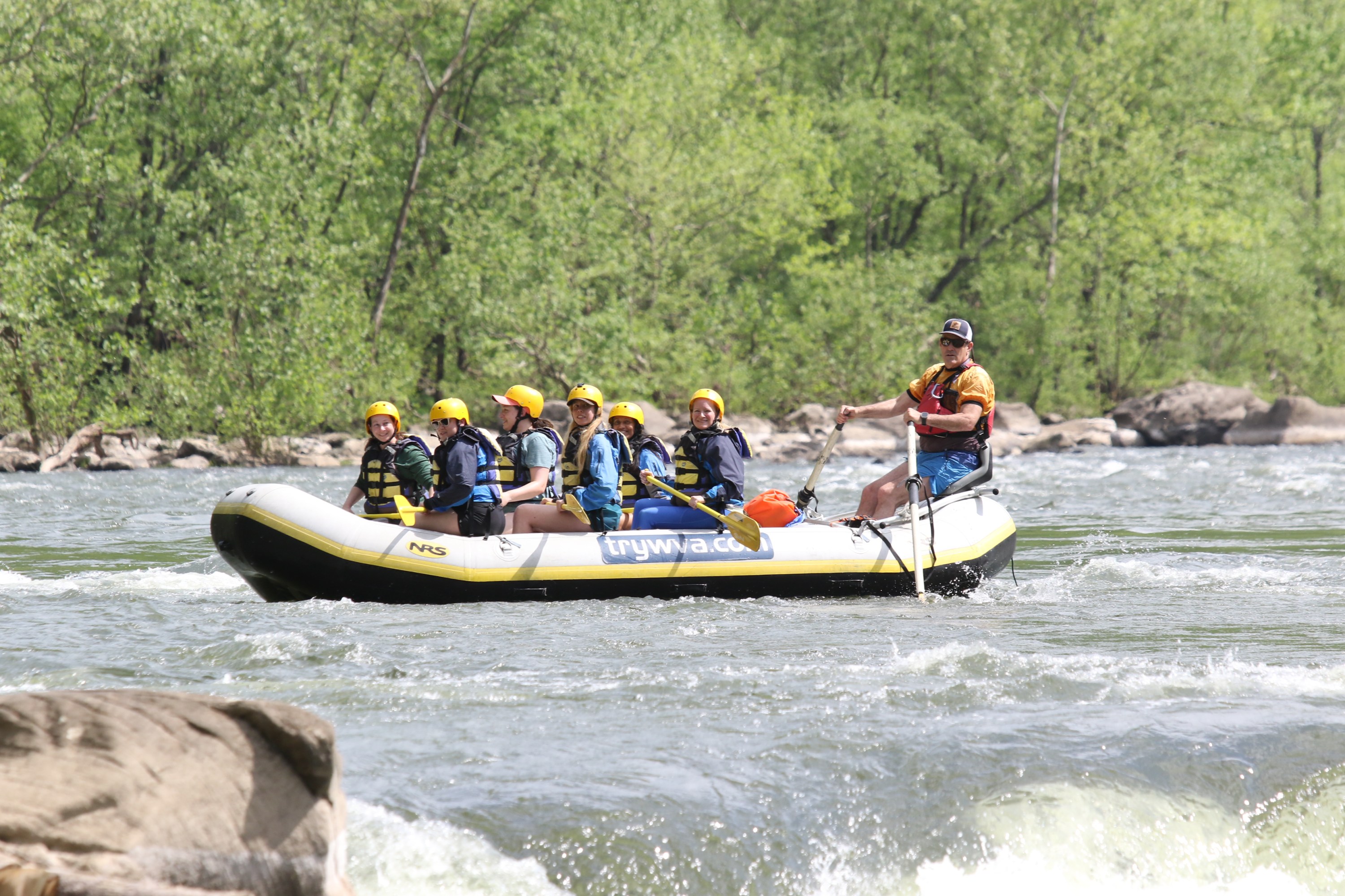 People in helmets rafting on a river surrounded by trees.