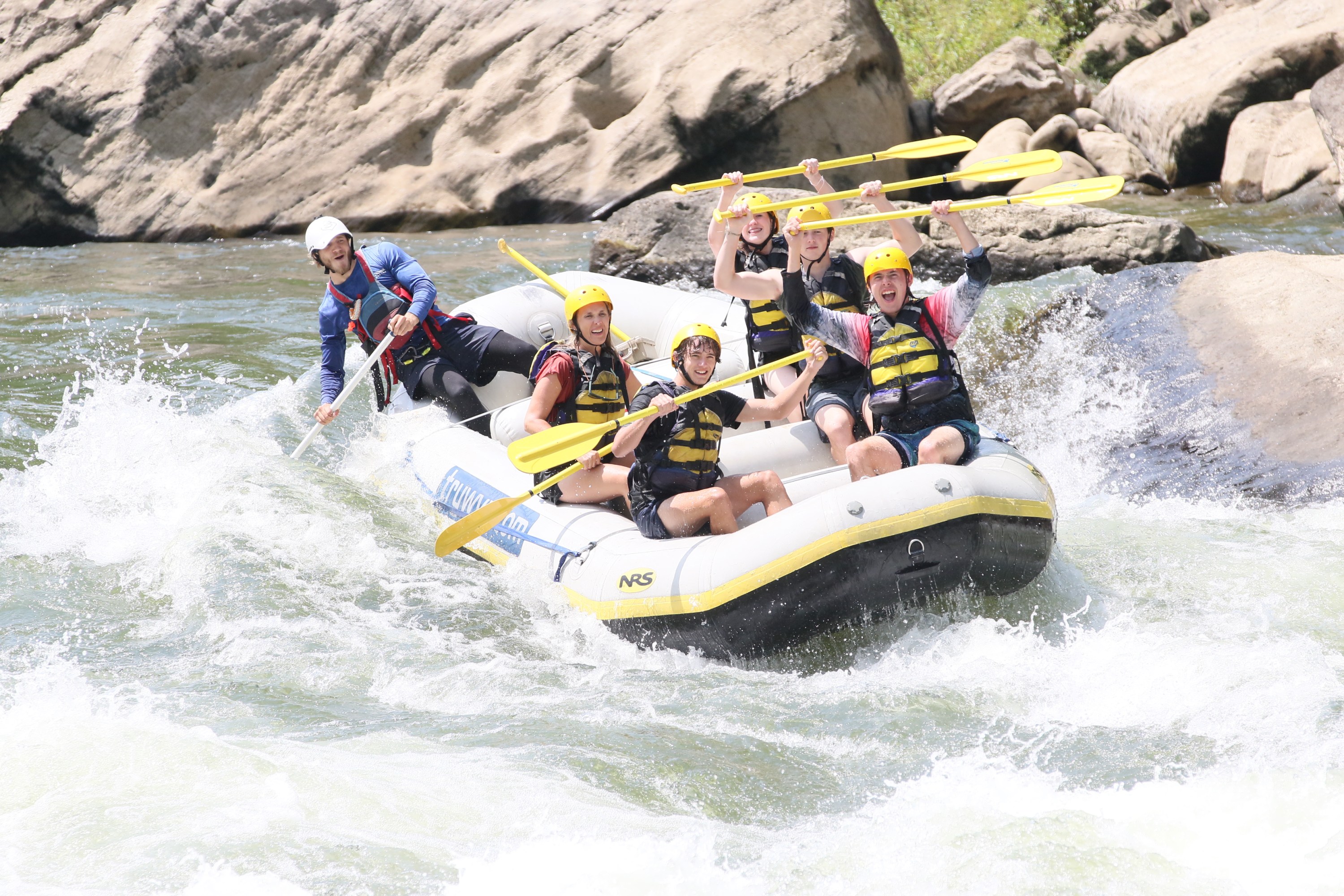 Group rafting on rapids, holding paddles up, wearing helmets and vests.