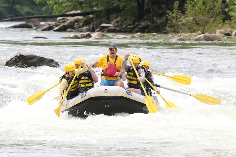 Group of people white water rafting on a river with yellow paddles.