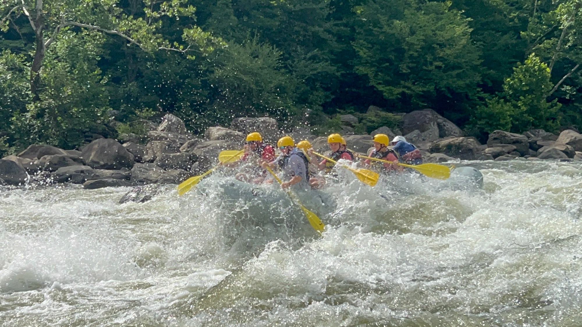 Group white-water rafting with yellow helmets in a rapid river.