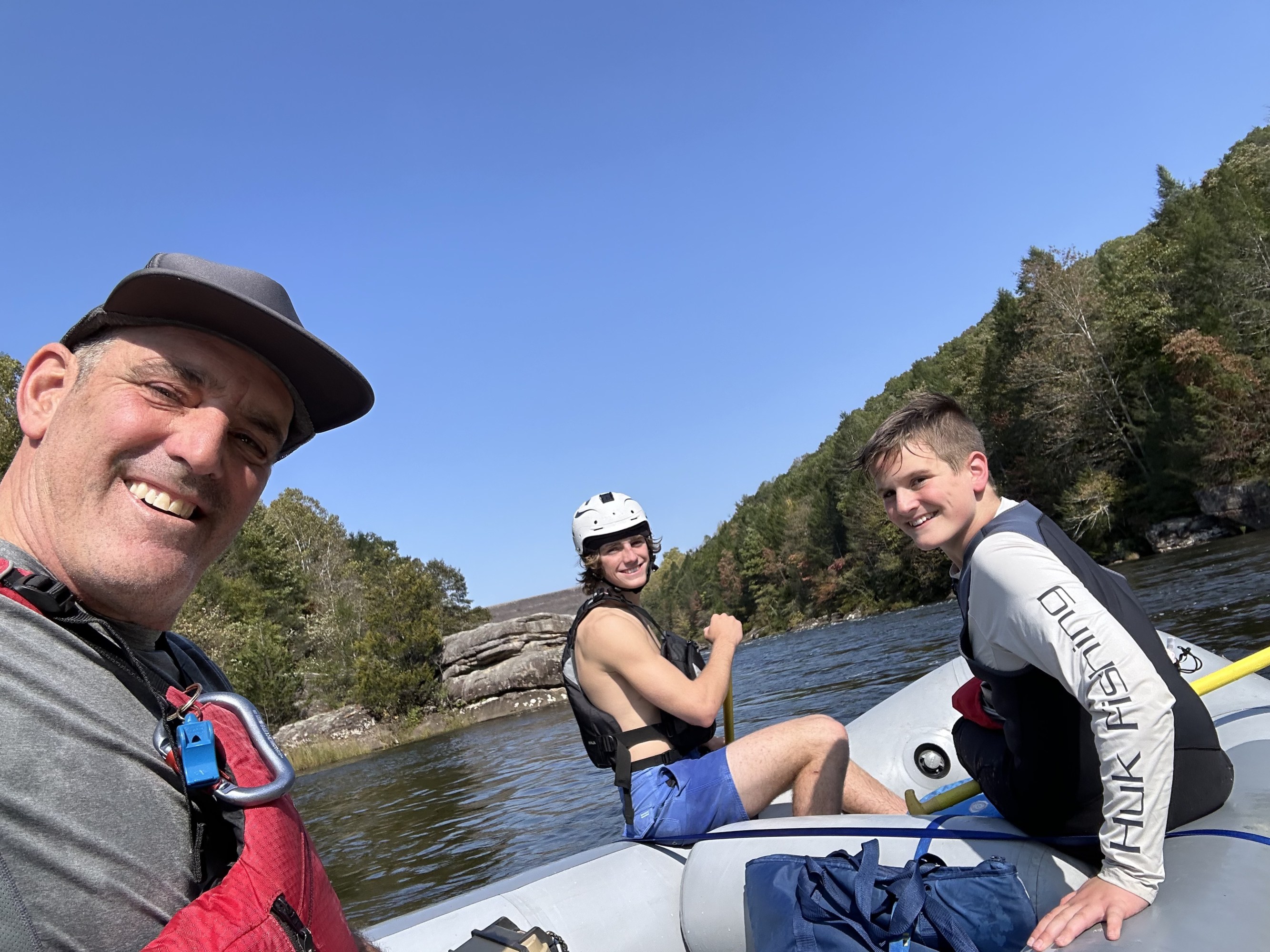 Three people in a raft on a river, wearing helmets and life jackets, with trees in the background.