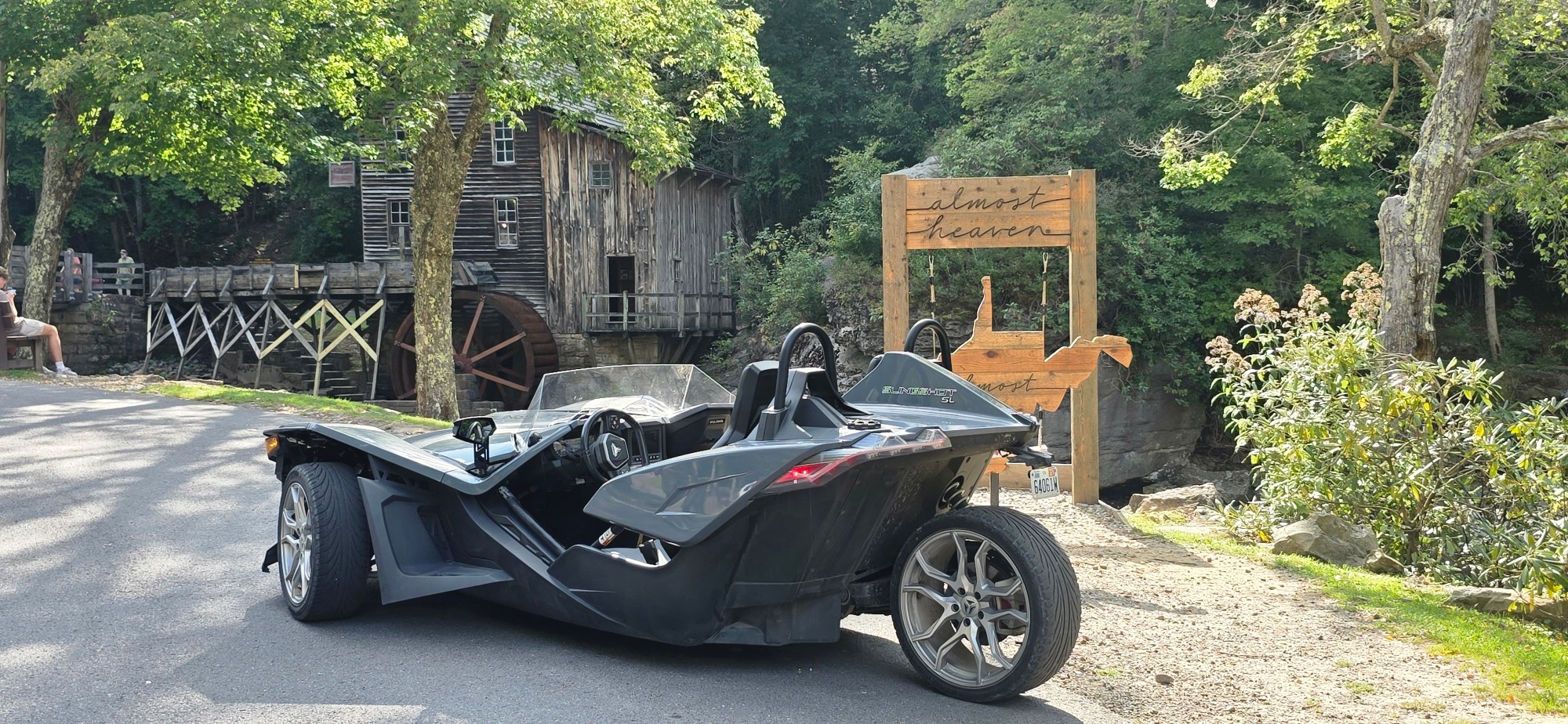 Three-wheeled vehicle parked near old mill with waterwheel and wooden sign in wooded area.