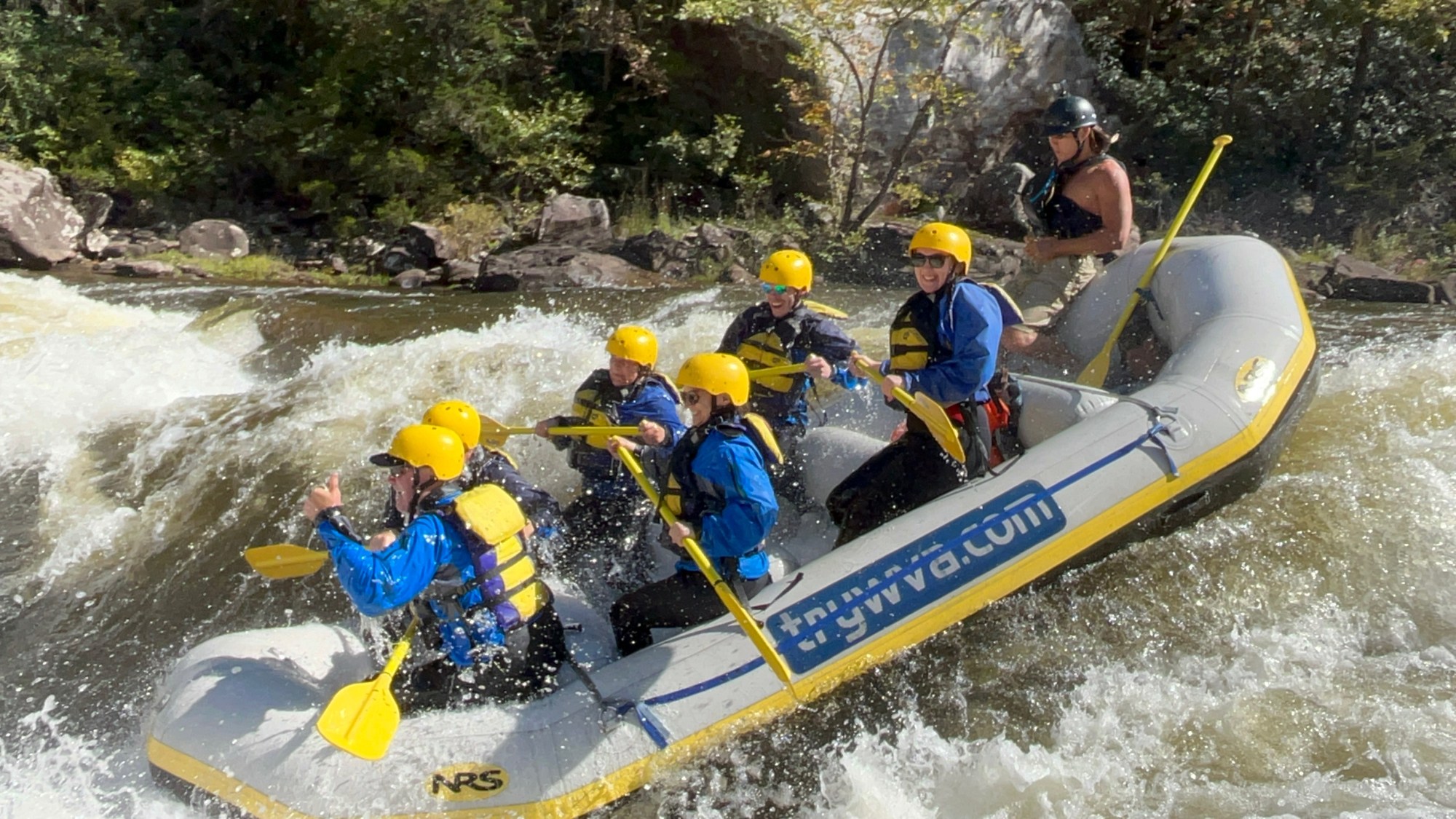 Group white water rafting in a raft with helmets and paddles on a river.