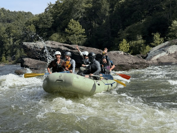 Group white water rafting on a river with helmets and paddles.