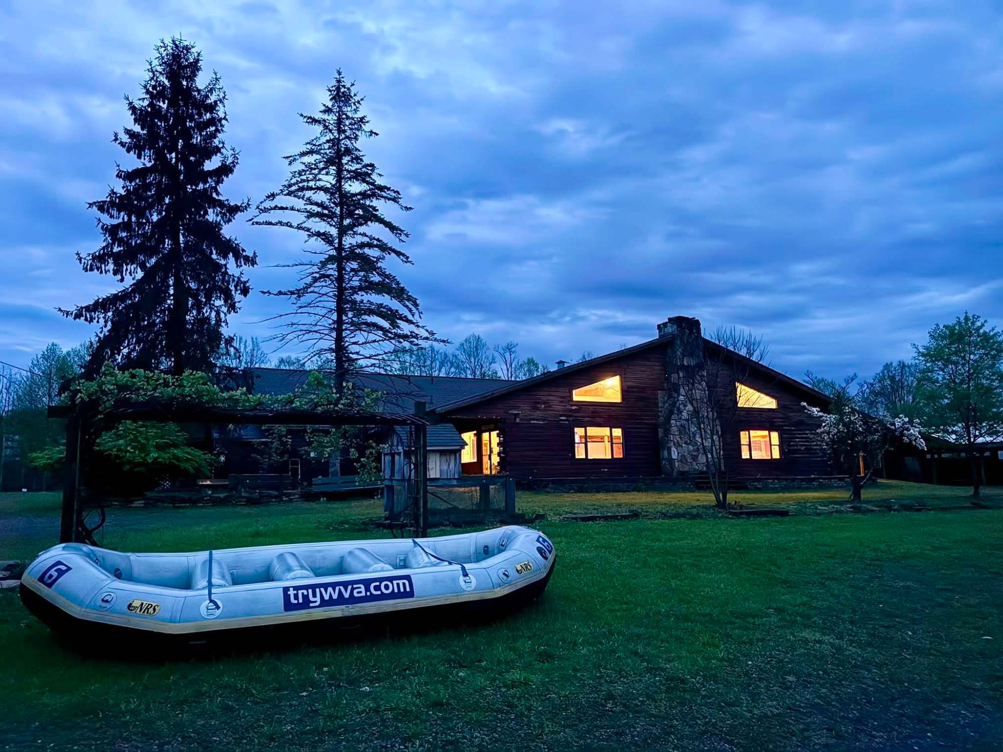 Inflatable raft on lawn near rustic cabin at dusk, pine trees in background.