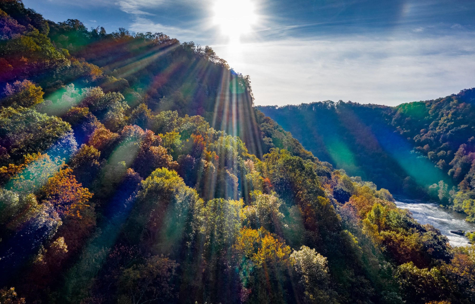 Sunlight beams over a forested valley with colorful autumn foliage and a river below.