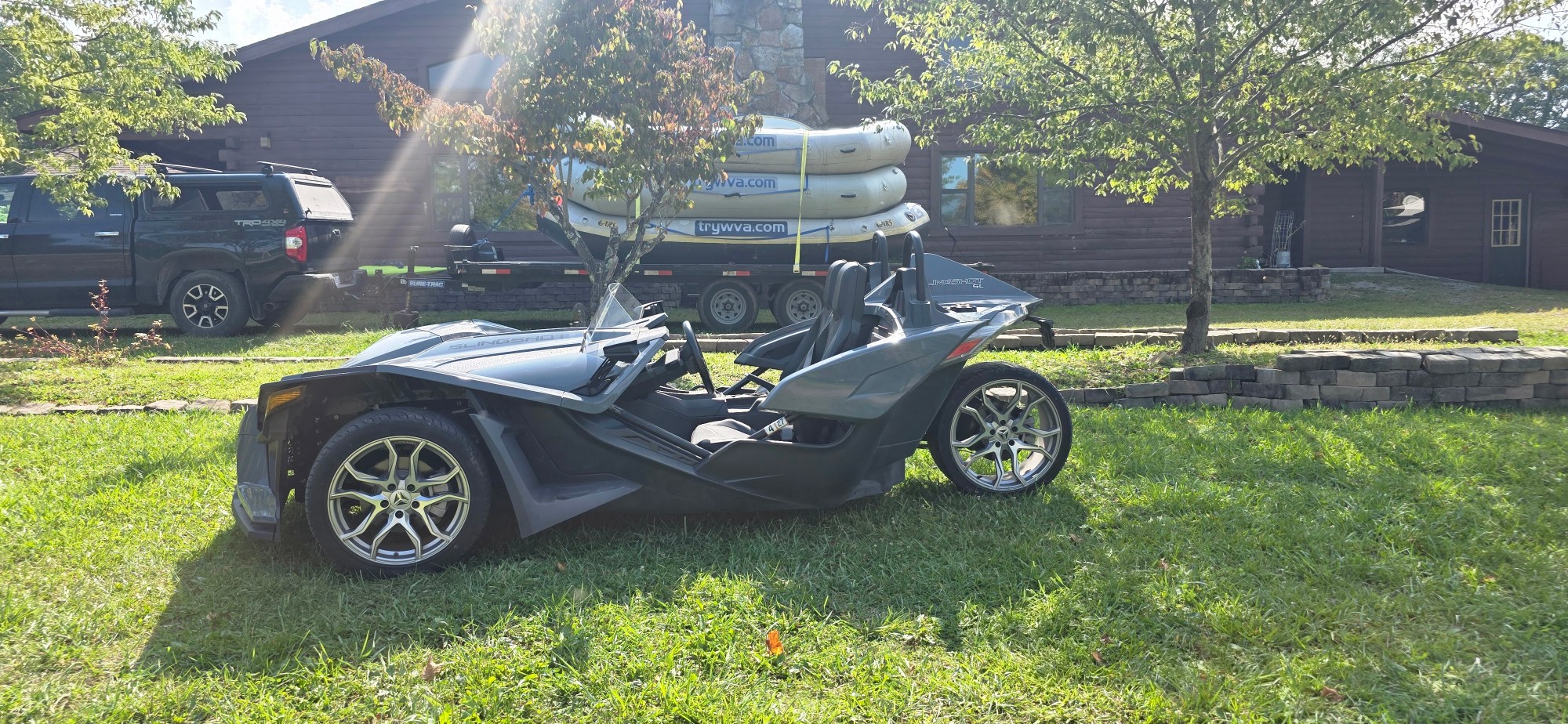 Three-wheeled vehicle parked on grass with rafts on truck in background.