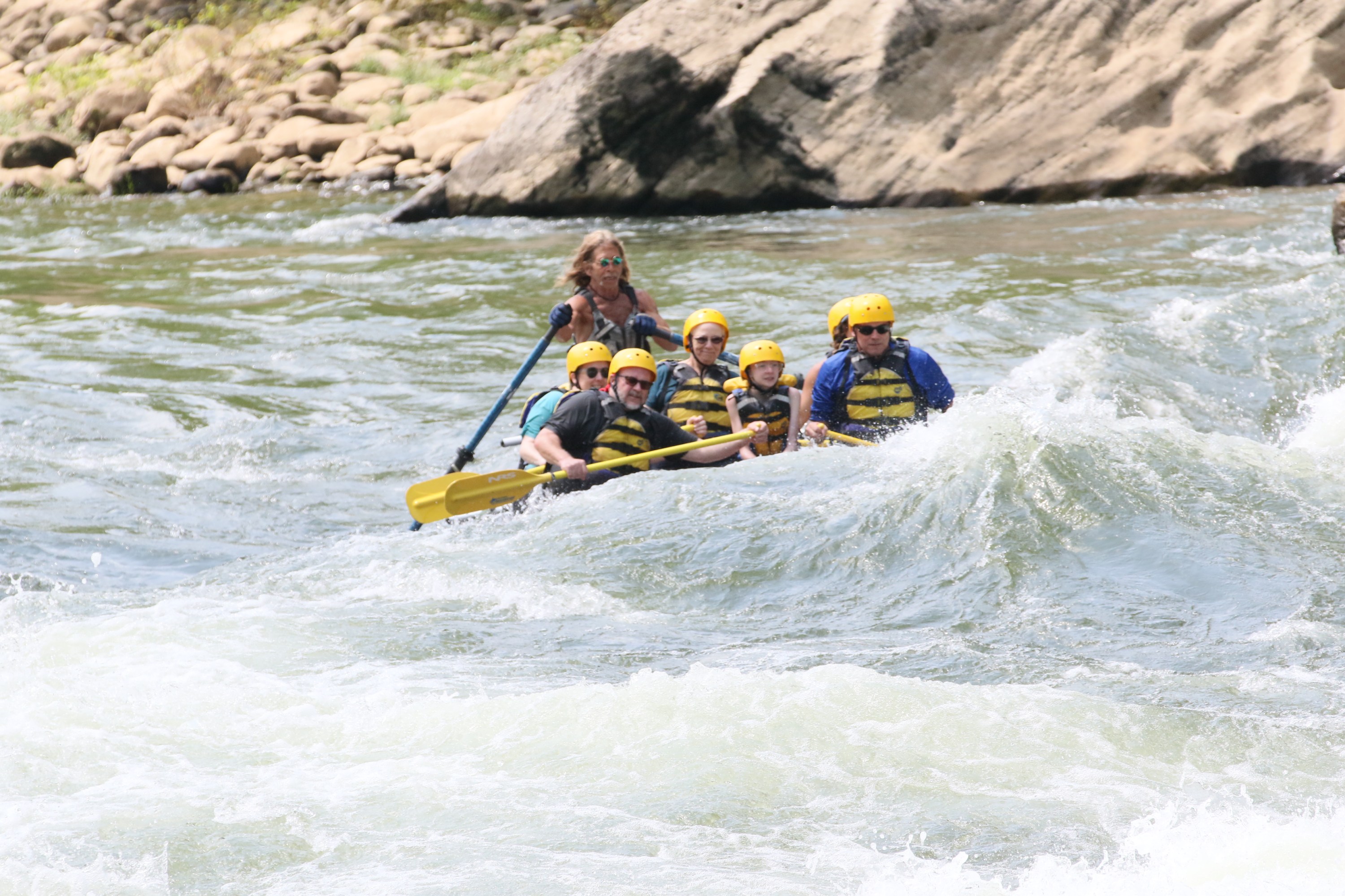 Group white-water rafting with helmets and life vests on a rocky river.